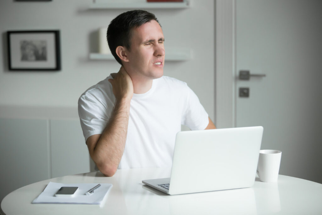 Young man with his hands at his neck pain, sitting at the white desk near the laptop, looking aside, home interior. Neck pain pose. Business concept photo, lifestyle